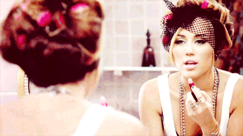 Miley Cyrus com redinha no cabelo, passando gloss labial frente a espelho com regata branca.
