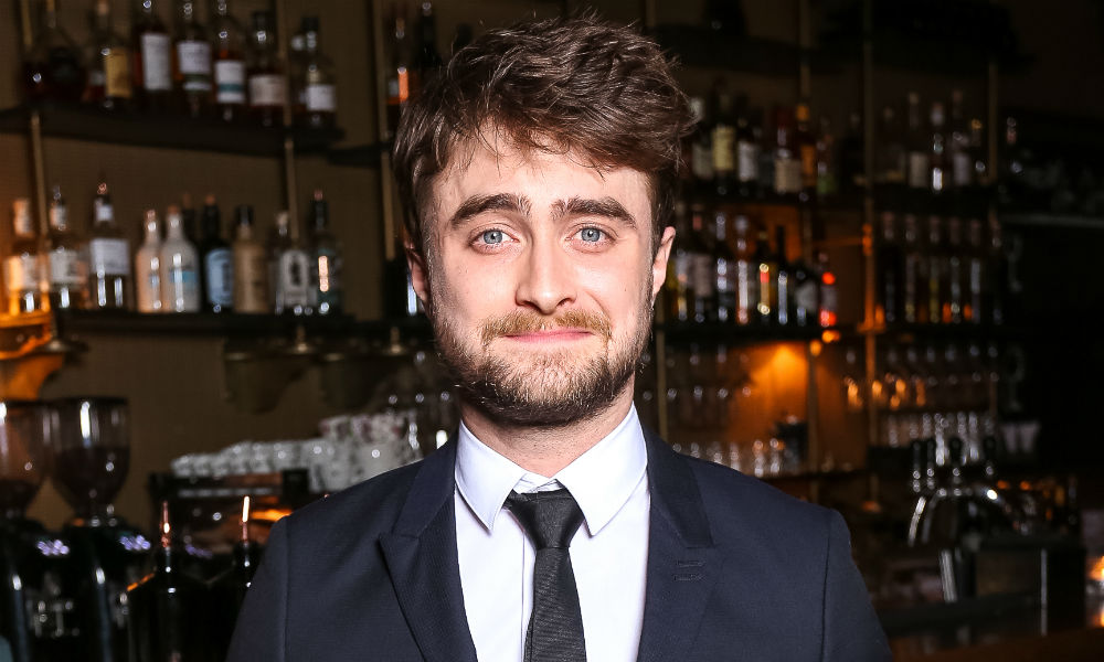 Daniel Radcliffe sorrindo levemente para direção da câmera usando terno preto e gravata preta com uma camisa branca com uma parede de bedidas de fundo
