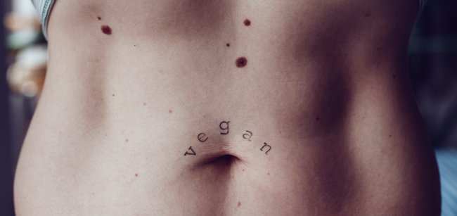 Foto da barriga de uma mulher branca e magra; em cima do umbigo, tem uma tatuagem com o escrito: vegan