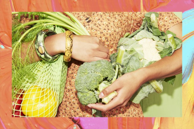 Foto dos braços de uma mulher voltando da feira. Ela veste um vestido laranja e está segurando várias verduras e muitos legumes
