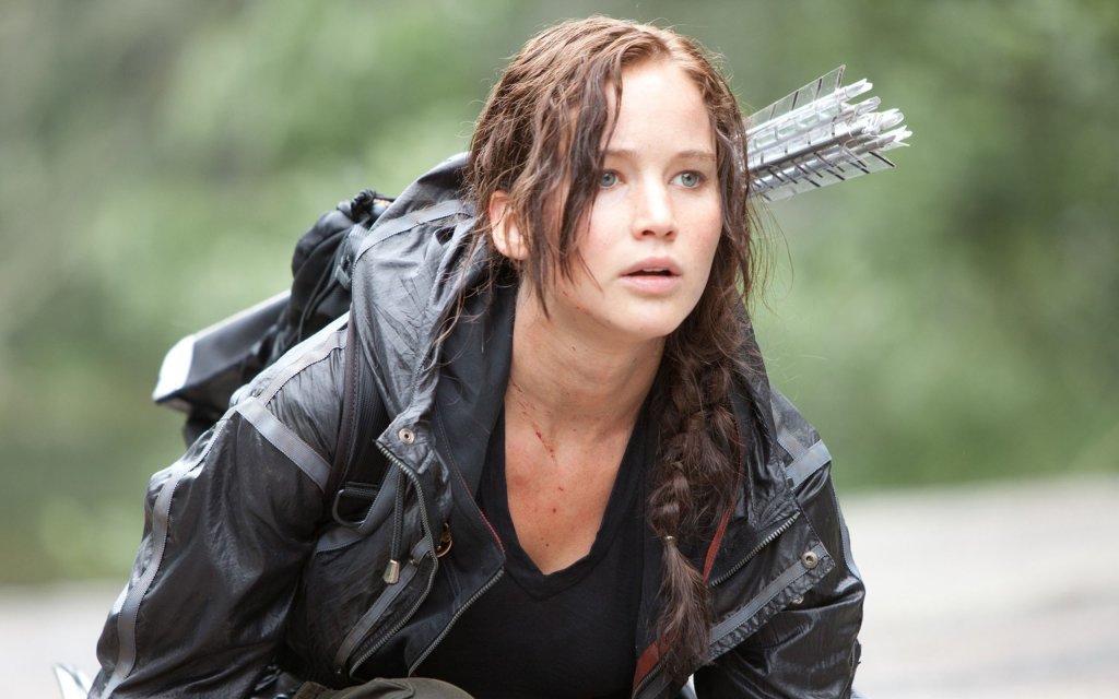 Katniss abaixada em cena de Jogos Vorazes com o arco e as flechas sendo carregadas nas costas; ela está com uma expressão de atenção