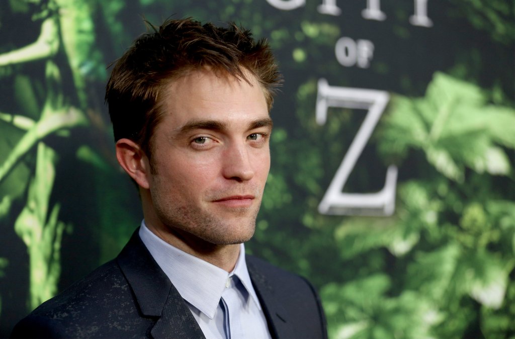 Robert Pattinson posando para foto em evento; o ator sorri levemente em um terno preto com camisa azul clara