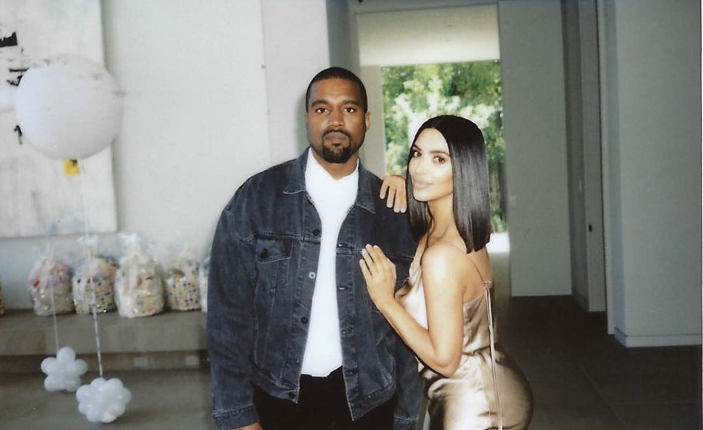 Kim e Kanye aparecem posando lado a lado. Ela está com um vestido bronze e ele de jaqueta jeans.