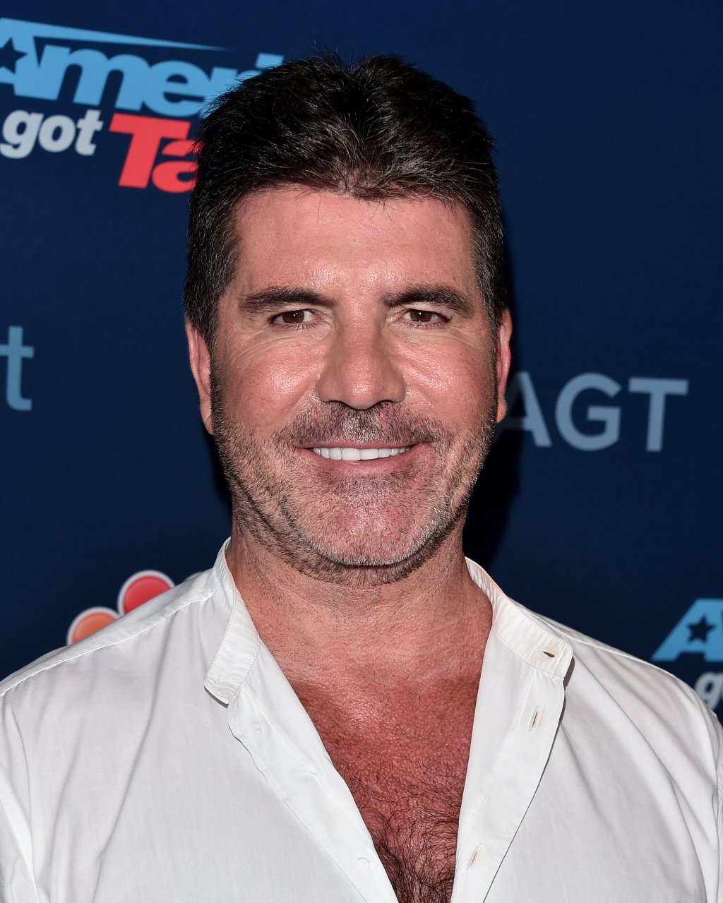 Simon Cowell com blusa branca sorrindo em fundo azul