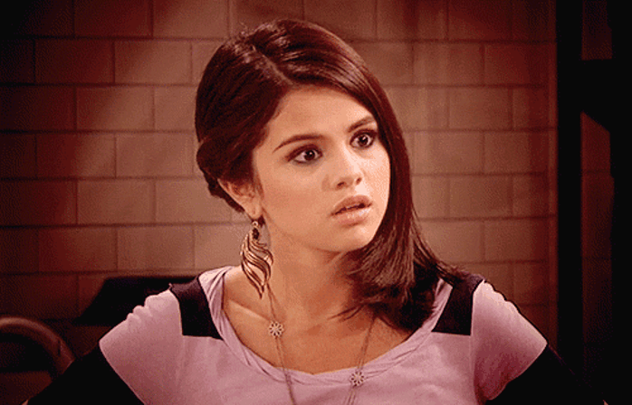 Selena Gomez com expressão de surpresa usando blusa rosa detalhe em preto.
