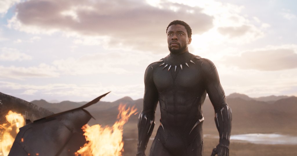 Chadwick Boseman caracterizado como Pantera Negra; ele está em uma cena do filme olhando para frente no meio de destroços que estão pegando fogo