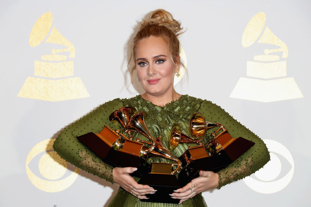 Adele em tapete vermelho com vestido verde e cabelo preso exibindo os cinco prêmios que ganhou no Grammy Awards 2017