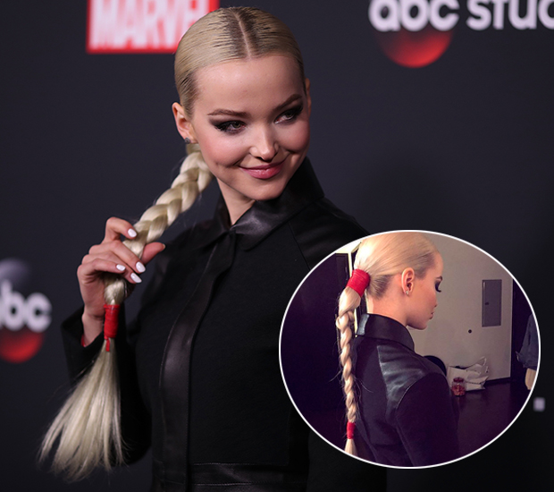 Dove Cameron usando look preto e segurando seu cabelo, que está preso em um rabo baixo com trança.