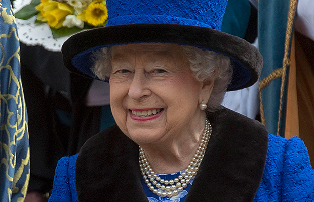 Rainha Elizabeth II usando joias de pérolas e um conjunto azul marinho com detalhes em preto, chapéu das mesmas cores, sorrindo abertamente para foto