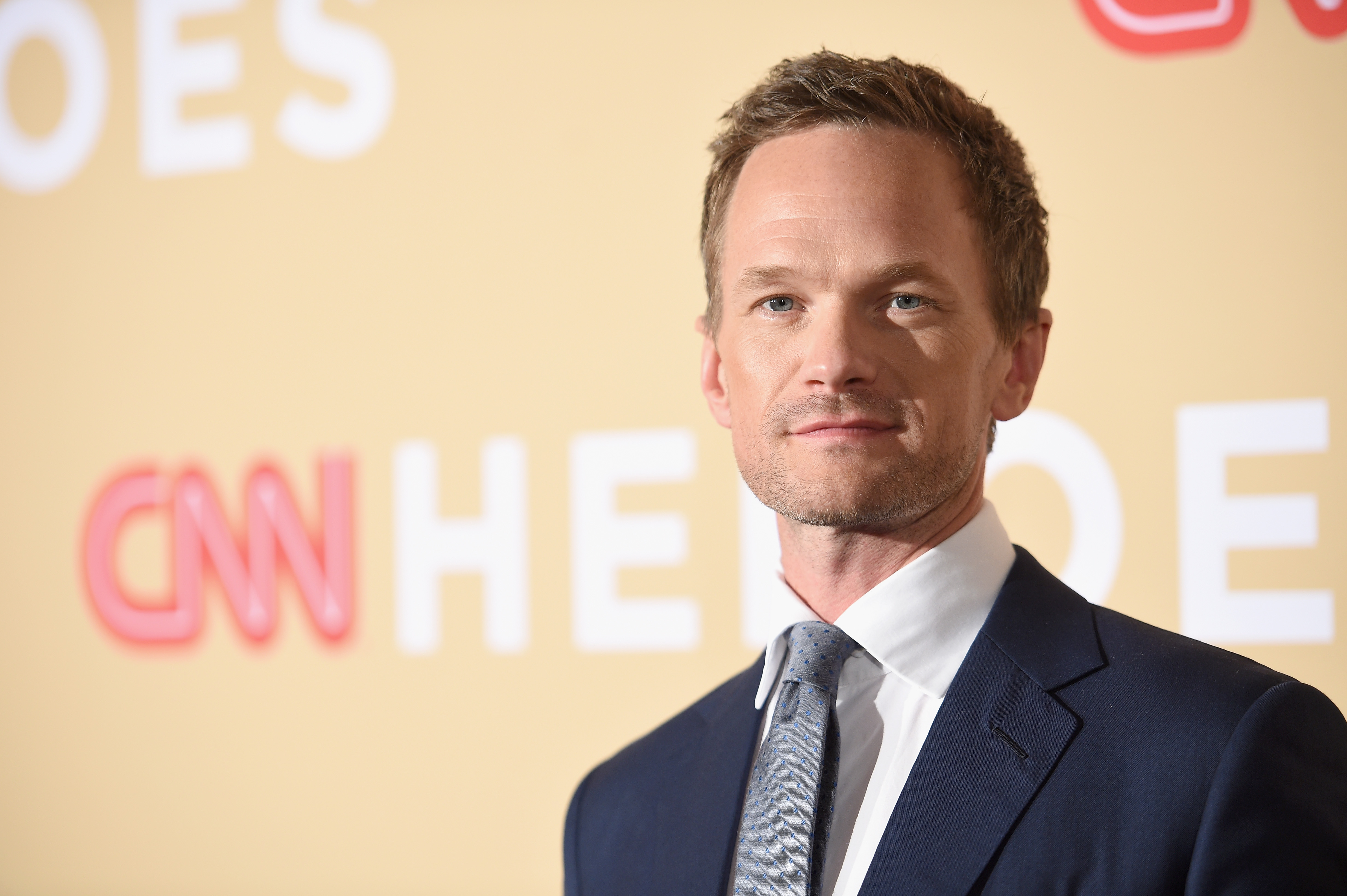 Neil Patrick Harris posando para foto no carpet do CNN Heroes 2015; ele usa um terno escuro com camisa branca e gravata cinza sorrindo levemente