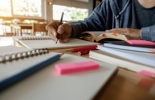 na imagem vemos uma mesa de estudo, com cadernos post-its e canetas. Ao fundo, tem um pessoa, que só aparece a mão escrevendo no caderno