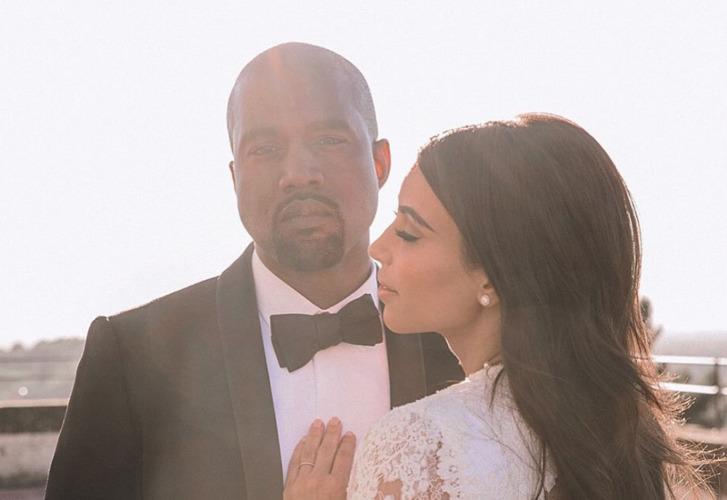 Foto de Kanye e Kim em seu casamento. O cantor aparece olhando para câmera com terno preto e camisa branca. A empresária está de lado com cabelos pretos e vestido de noiva.