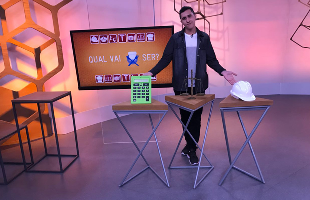 programa-como-serc3a1 A imagem mostra um homem com camisa jeans e calça preta, em um estúdio de tv, em frente a 3 mesas, cada uma com um acessório diferente me cima: uma calculadora, uma balança em estilo antigo, com dois pratos e um capacete de proteção na cor branca. Atrás dele há uma tv com um fundo laranja escrito: Qual vai ser em letras brancas.