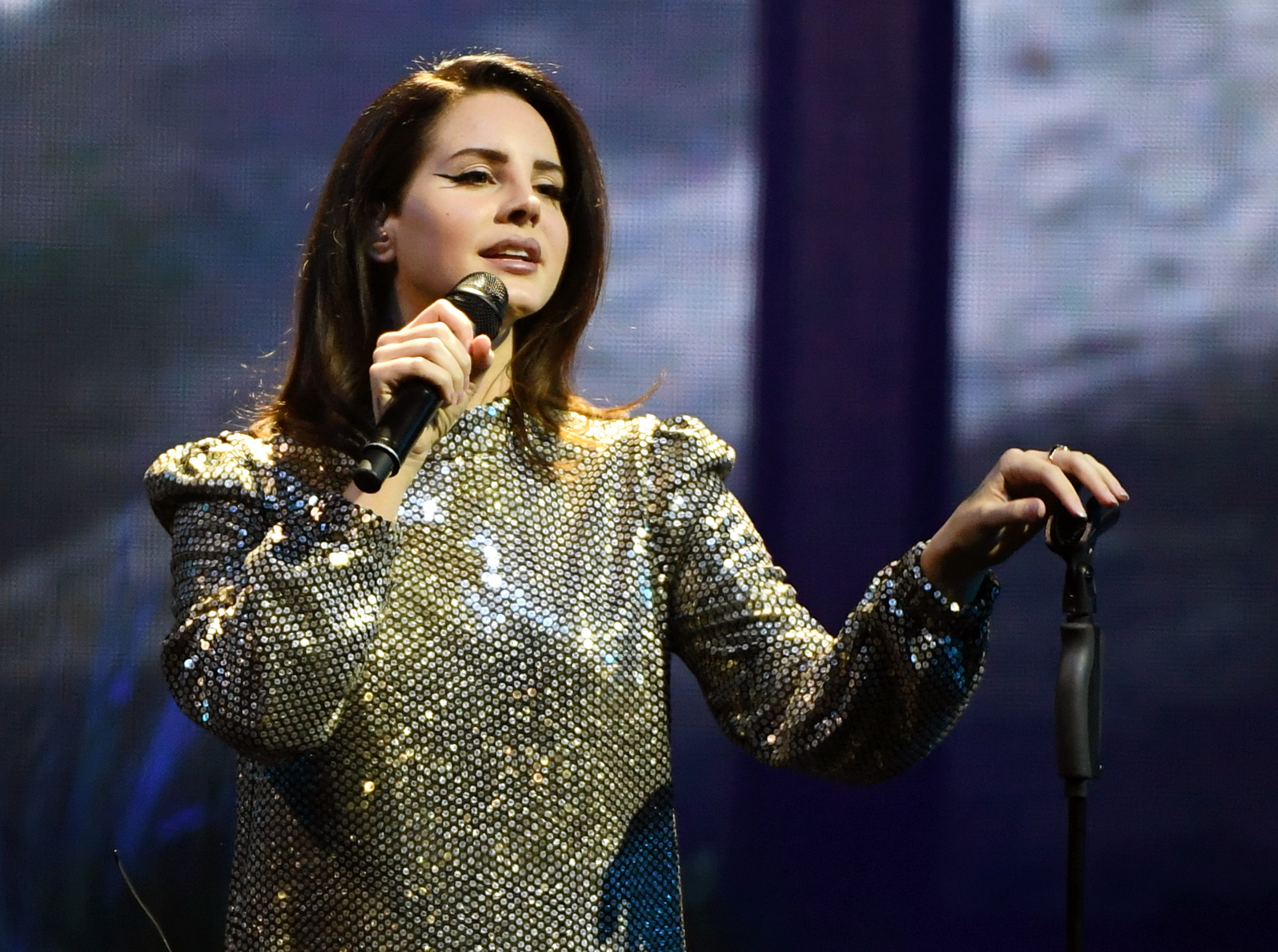 Lana Del Rey cantando no show em Mandalay Bay, em Las Vegas; ela usa uma roupa brilhante e olha para o público segurando o microfone em uma das mãos