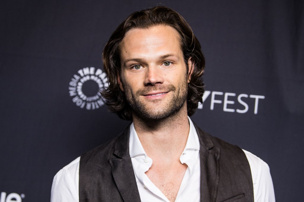 Jared Padalecki posando para foto em carpet com camisa branca e colete preto; o ator sorri levemente