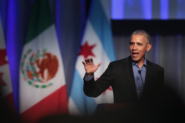barack-obama Barack Obama com a mão levantada enquanto fala em discurso; ele usa terno preto com uma camisa azul