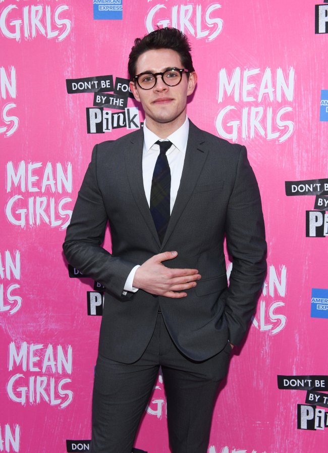 Casey Cott posando para carpet de Mean Girls; ele usa óculos com terno cinza escuro com camisa branca e gravata que mescla preto e azul em estampa xadrez; o ator sorri levemente e está com uma das mãos na barriga
