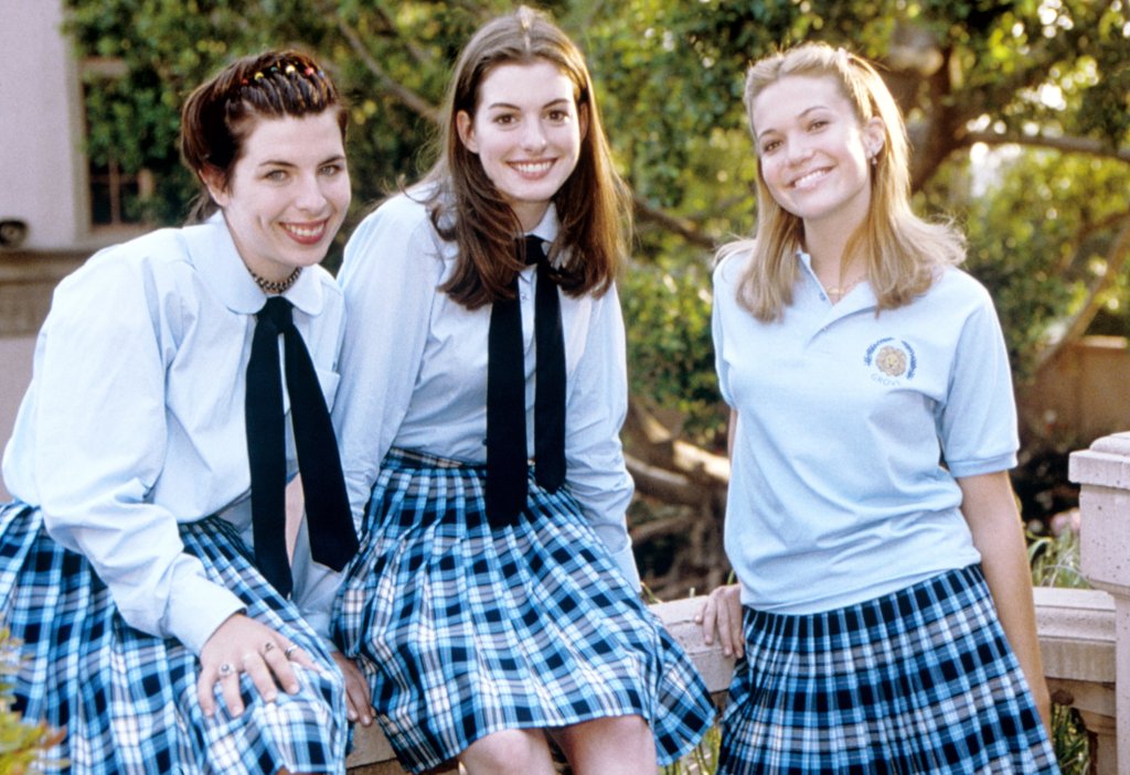 Três mulheres sorrindo com uniformes escolares que contam com saia azul quadriculada com preto, camisa azul e duas delas usam gravata enquanto outra usa uma camiseta estilo polo