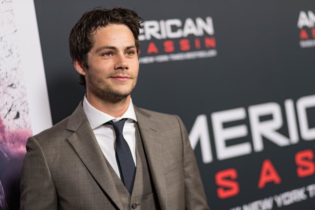 Dylan O'Brien posando para foto em carpet de evento com terno marrom, camisa branca e gravata preta; o ator sorri levemente