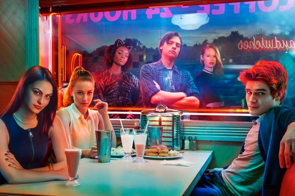 Foto de divulgação de Riverdale; Camila Mendes, Lili Reinhart e KJ Apa estão dentro da lanchonete sentados na mesa e o restante do elenco aparece posando do lado de fora sendo vistos pelo vidro da janela;o ambiente está iluminado por luzes coloridas e neon