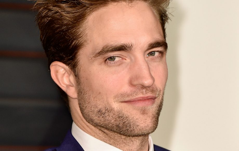 Foto com close no rosto de Robert Pattinson; o ator sorri levemente com barba curta enquanto olha para o lado