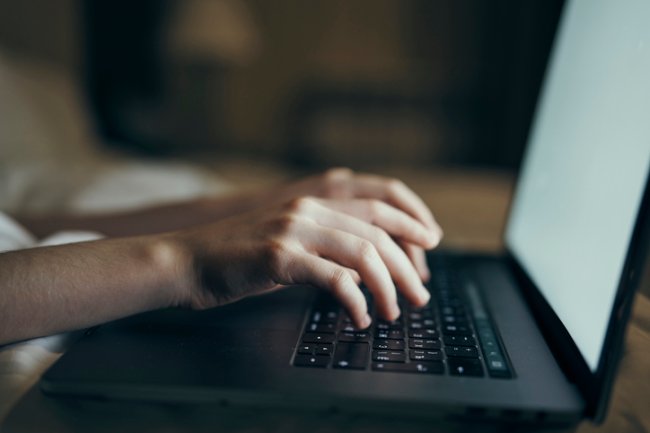 A imagem mostra duas mãos de uma pessoa branca digitando em um computador preto, com a tela ligada. O fundo da imagem é desfocado.
