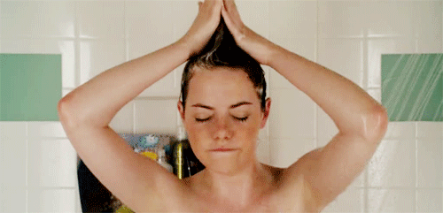 Gif da Emma Stone no banho lavando o cabelo, cantando e segurando o cabelo no alto com as duas mãos.