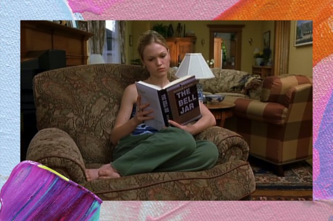 Uma mulher branca e loira, por volta dos 20 anos, sentada numa poltrona marrom e lendo um livro