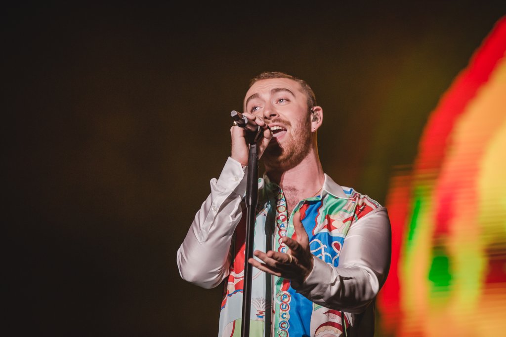 Sam Smith no palco do Lollapalooza Brasil; o cantor segura a base do microfone com uma mão e estende a outra na direção do público; ele olha para frente e luzes coloridas iluminam o fundo do palco