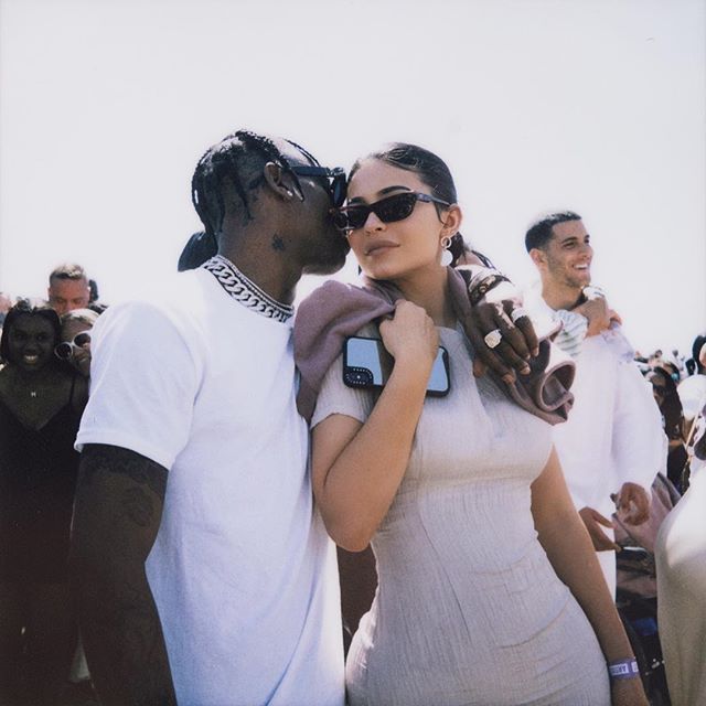 Kylie Jenner e Travis Scott posando para foto; Travis está usando uma camiseta branca enquanto dá um beijo na bochecha de Kylie com o braço ao redor do pescoço da empresária que usa um vestido claro com óculos de sol e sorri levemente