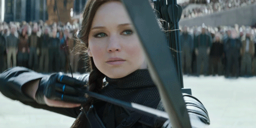 Moving image of Katniss pointing a bow and arrow with a serious expression;  she has her hair in a braid and wears gloves
