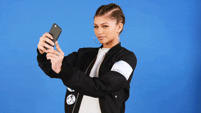 Gif da Zendaya usando camiseta branca e jaqueta preta e brincos de argola. Ela está segurando um celular com as duas mãos para tirar uma selfie.