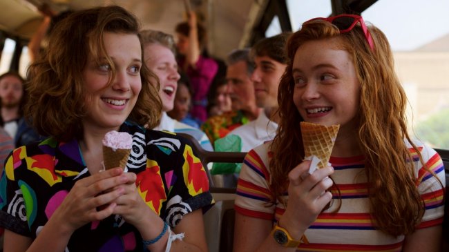 Millie Bobby Brown e Sadie Sink em cena de Stranger Things; elas estão sentadas em um ônibus olhando uma para outra segurandos sorvete e sorrindo