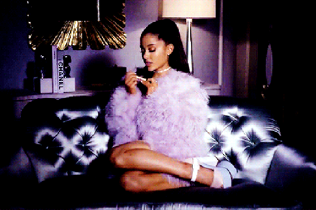 Ariana Grande lixando as unhas em gif que faz close no rosto dela, Ariana usa casaco de pelinhos lilás, e sapato da mesma cor. Sua expressão é séria.