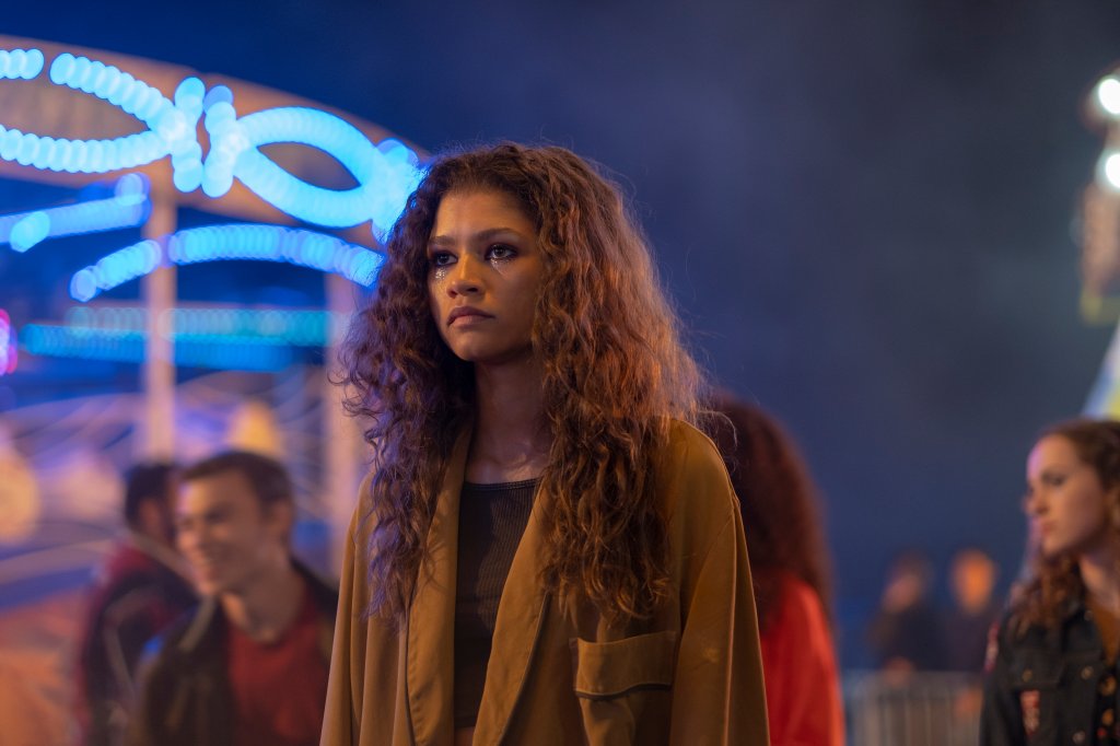 Zendaya como Rue em Euphoria; ela está parada no meio da multidão com uma expressão triste; a atriz usa um casaco marrom com blusa preta e maquiagem colorida