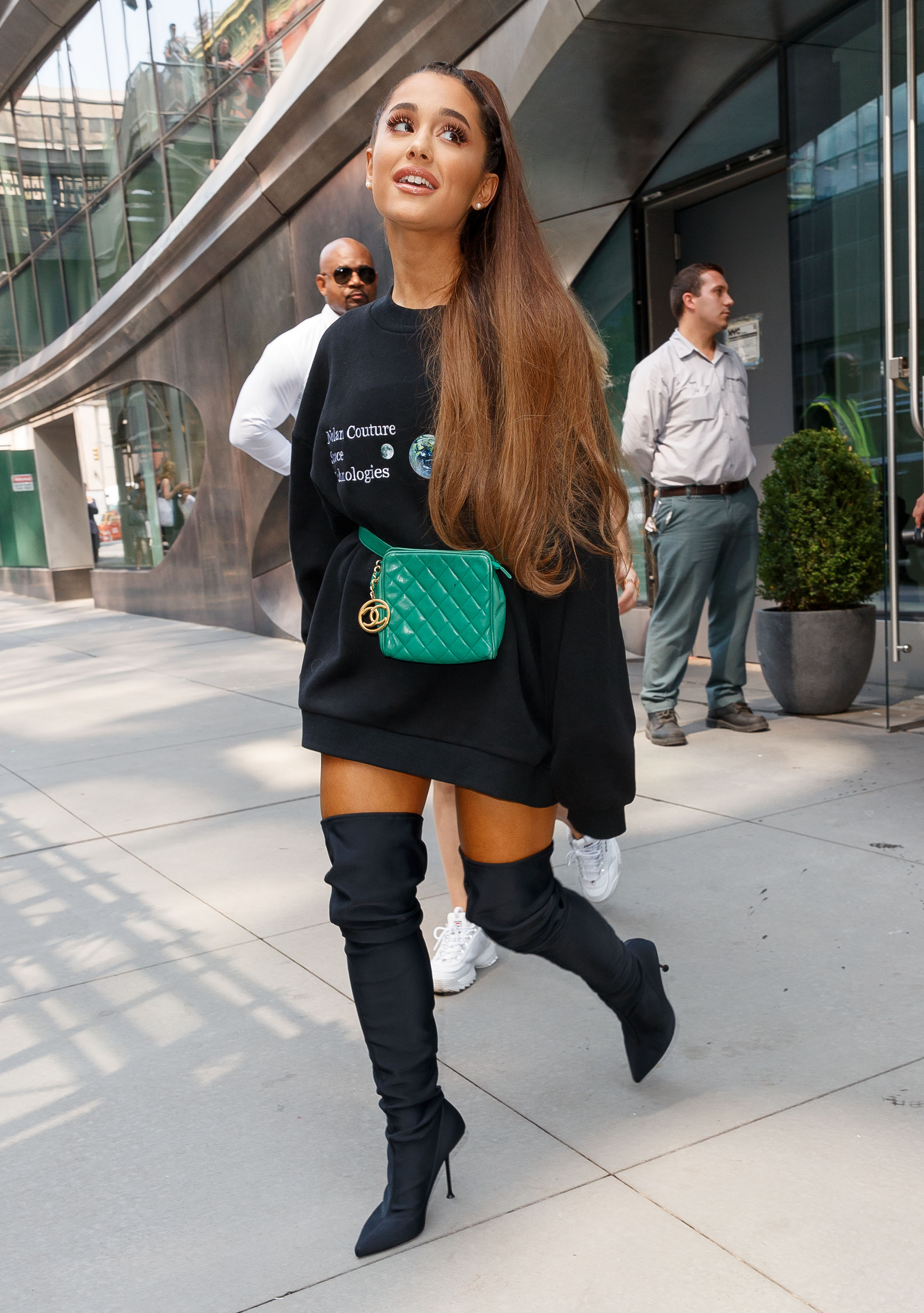 Ariana Grande usando moletom preto oversized com pochete verde e bota over the knee preta.