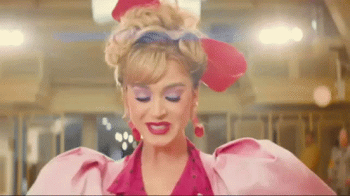 Gif da Katy Perry andando e mandando beijo com uma das mãos. Ela está usando roupa vermelha e rosa.