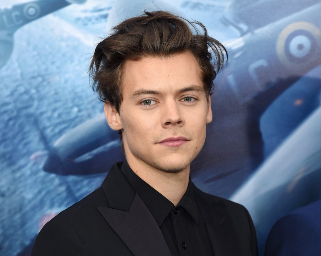 Harry Styles posando para foto sorrindo levemente; Ele está com terno e camisa na cor preta e o fundo é um backdrop em tons de azul