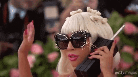 GIF da lady Gaga usando peruca loira chanel, óculos de sol escuros com strass, batom rosa e segurando um telefone na orelha enquanto faz bico com os lábios.