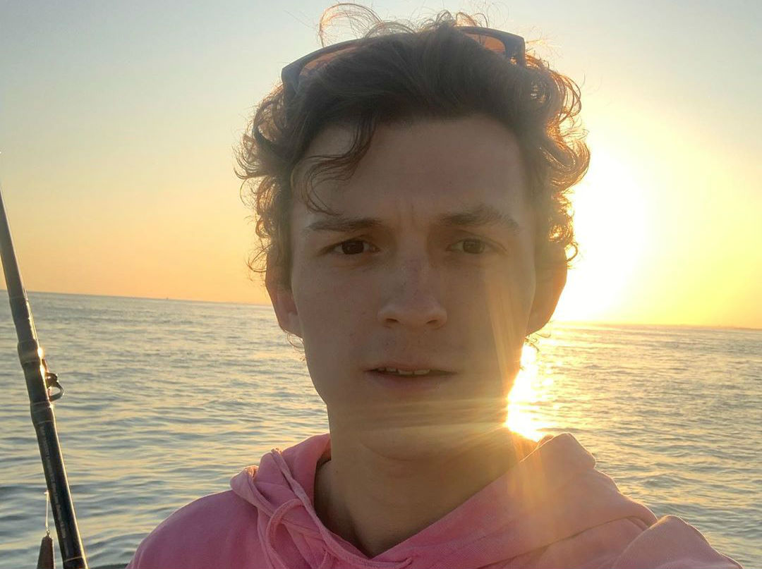 Selfie de Tom Holland no pôr-do-sol; ele usa um moletom rosa e é possível ver o mar no fundo da foto
