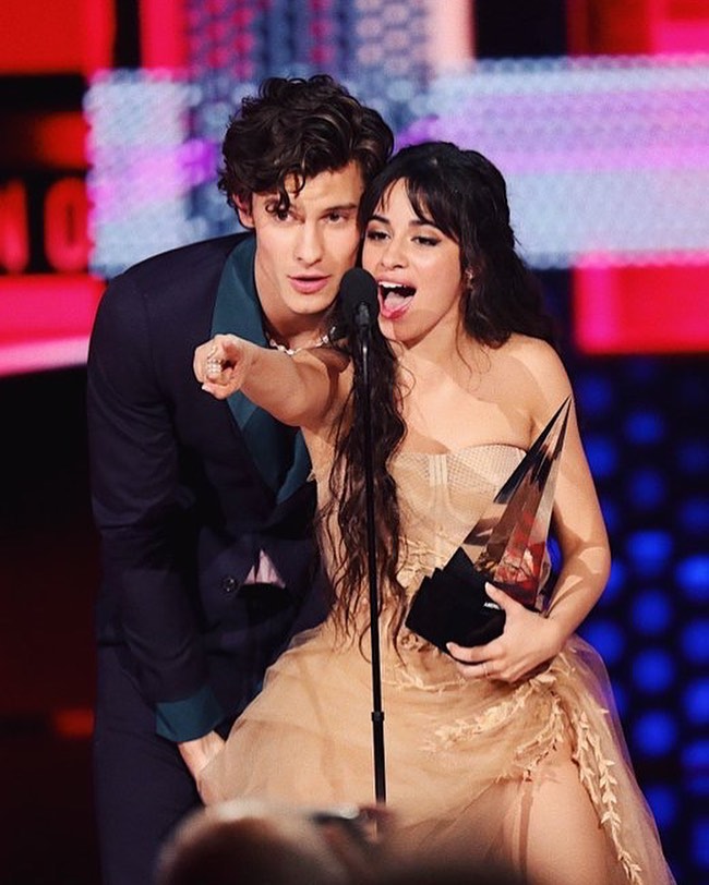 Camila Cabello e Shawn Mendes ganharam o prêmio de Colaboração do Ano no American Music Awards; Os dois estão no palco, Camila está segurando o prêmio e apontando para o público enquanto Shawn está abaixado com o queixo apoiado no ombro da cantora e olhando na direção em que ela aponta