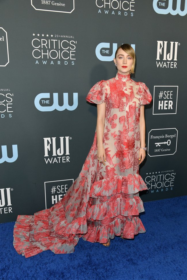 Saoirse Ronan com vestido todo romântico com detalhes vermelhos e babados; ela está com expressão neutra, cabelo preso e usa batom vermelho posando no carpet do Critics Choice Awards promovido pela CW e pela FIJI que aparecem com seus logos no backdrop ao fundo da imagem