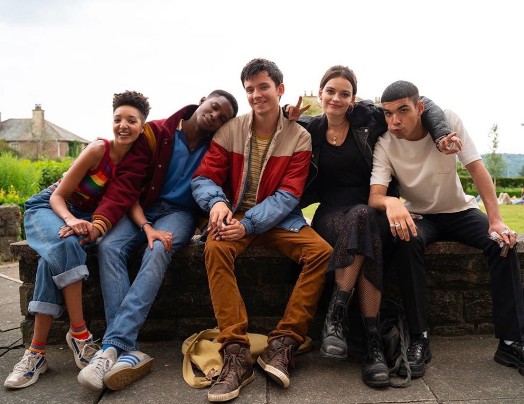 Elenco de Sex Education nas gravações da 2ª temporada, todos estão sentados e abraçados enquanto mostram sorrisos para a câmera