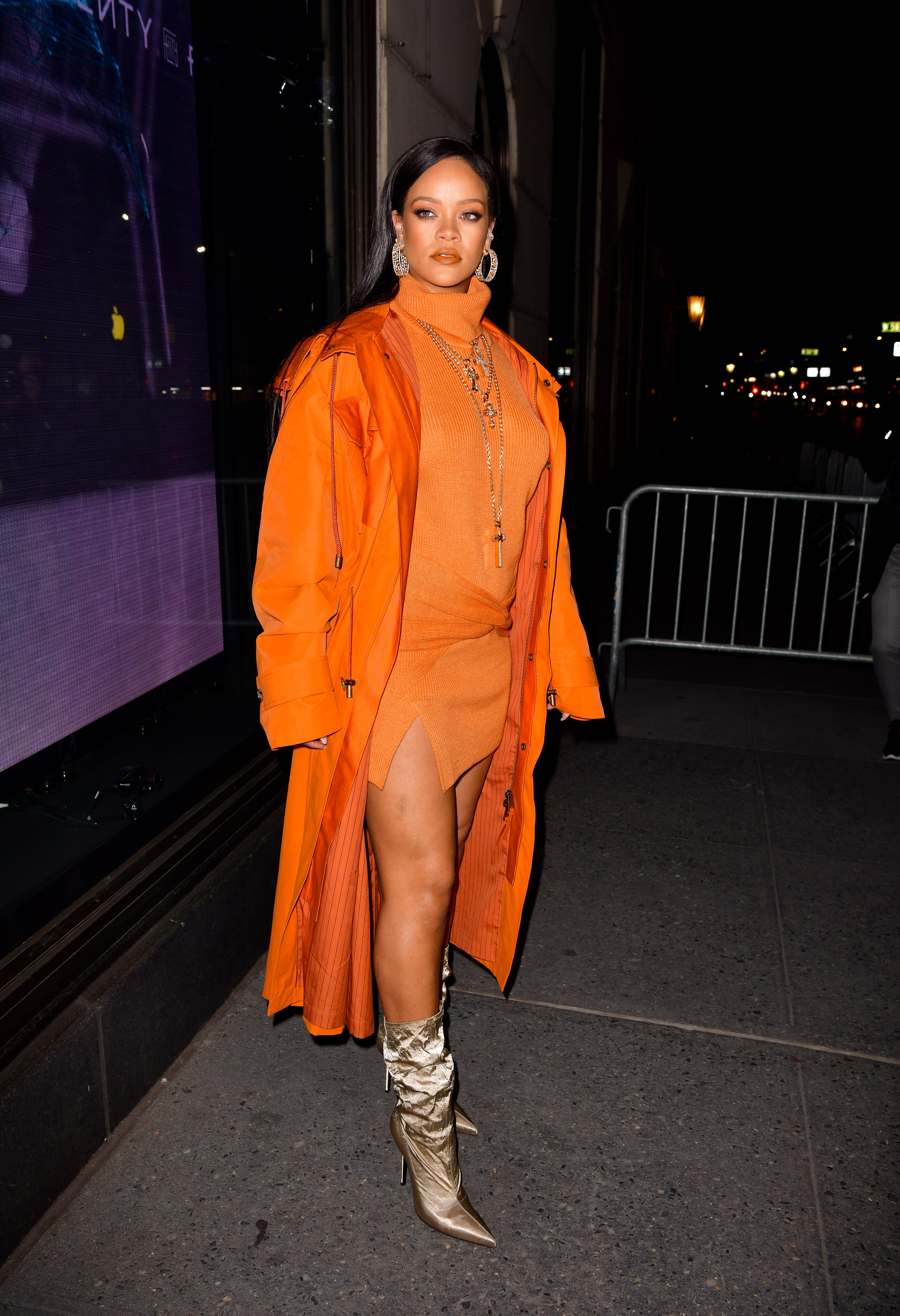 Rihanna usando vestido de gola alta laranja, sobretudo no mesmo tom de laranja e bota slouchy dourada. Ela está com vários colares dourados, olhando para o lado com expressão facial séria.
