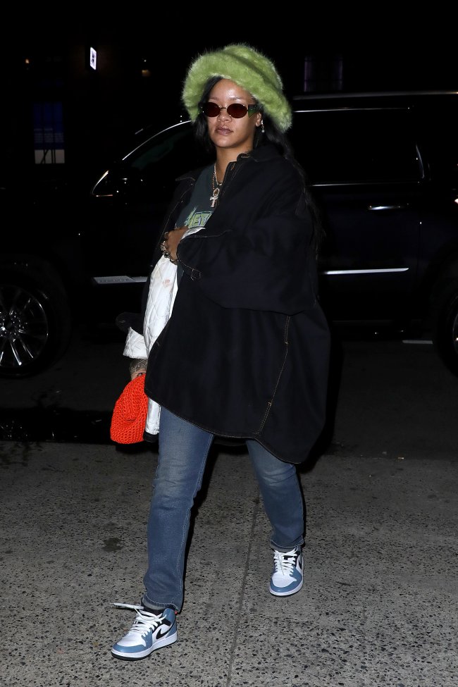 Rihanna usando bucket hat verde de pelinhos, casaco preto, calça jeans, tênis Nike Air Jordan azul e branco e óculos de sol, segurando uma bolsa laranja.