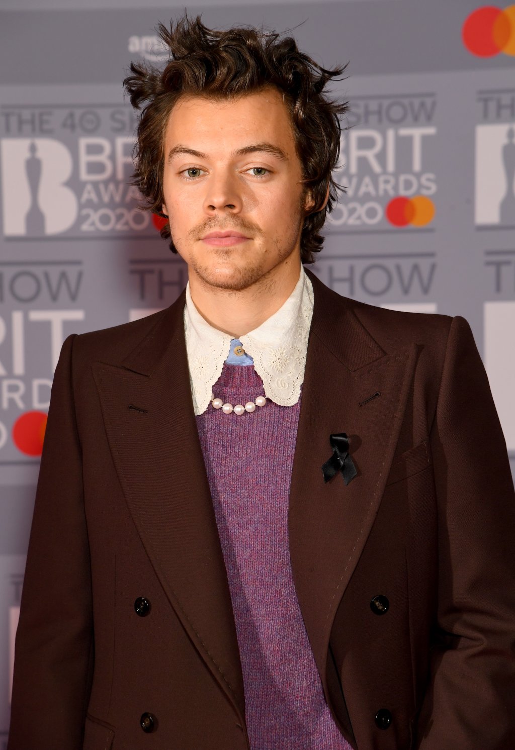 Harry Styles usando terninho da Gucci no BRIT Awards com expressão facial séria, suéter roxo e gola da camisa branca aparente