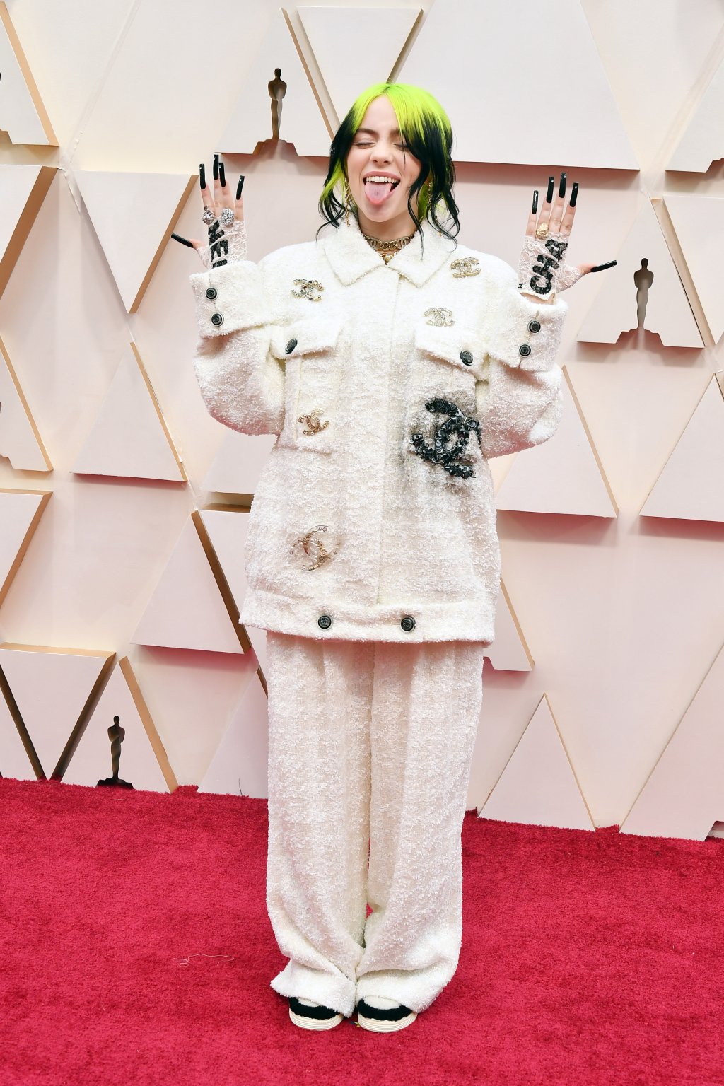 Foto da Billie Eilish no tapete vermelho do Oscar em 2020. Ela usa um conjunto da Chanel branco e tênis. Ela sorri e mostra a língua para a foto.