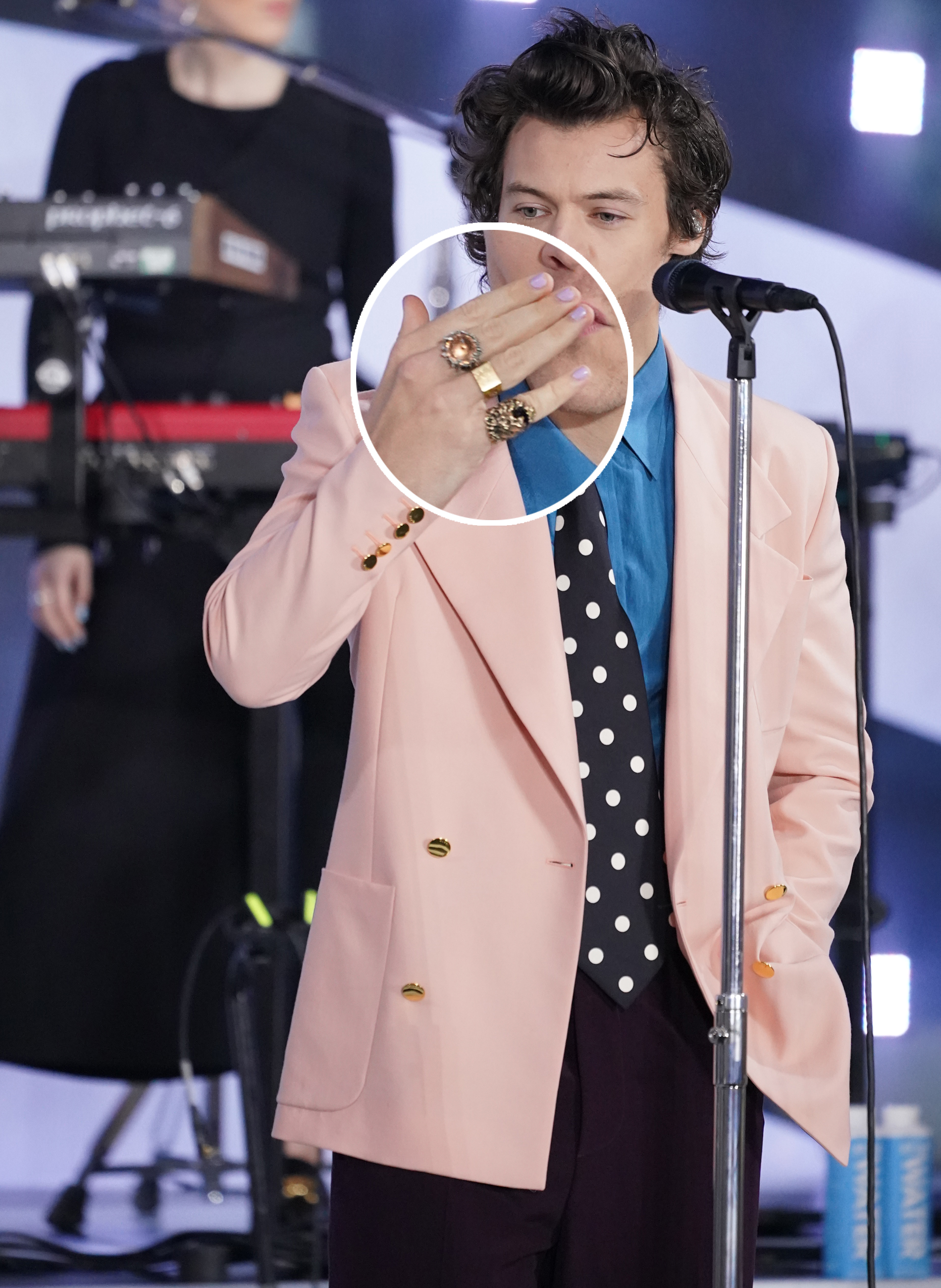 Harry Styles usando blazer rosa com gravata preta de poá, camisa azul e mix de anéis nos dedos. Ele está com a mão na boca mandando beijo para o público.