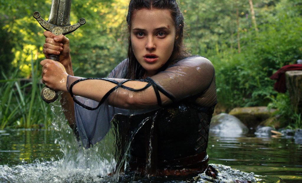 Katherine Langford em cena de Cursed; ela está dentro de um lago segurando uma espada com expressão séria