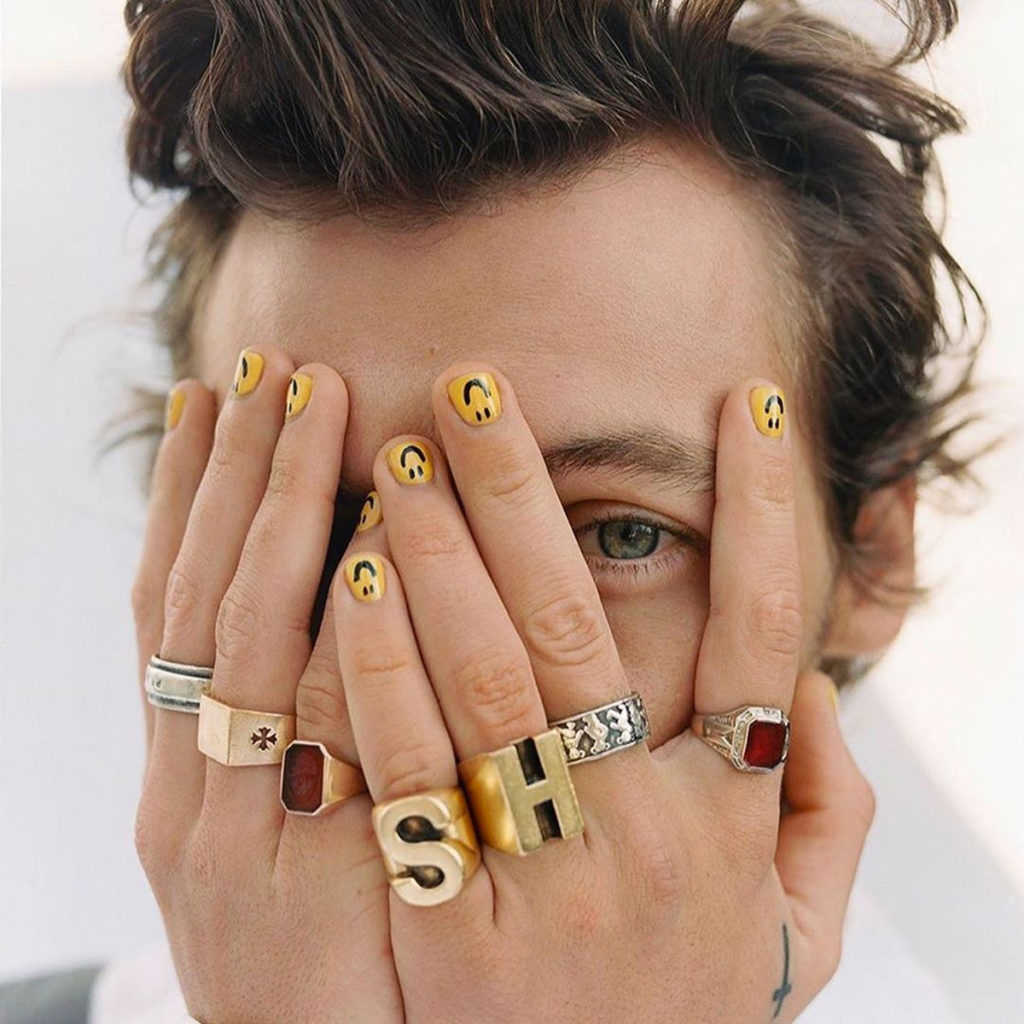 Harry Styles cobrindo o rosto com as duas mãos, que estão cheias de anéis diferentes e unhas pintadas de esmalte amarelo e carinha feliz.